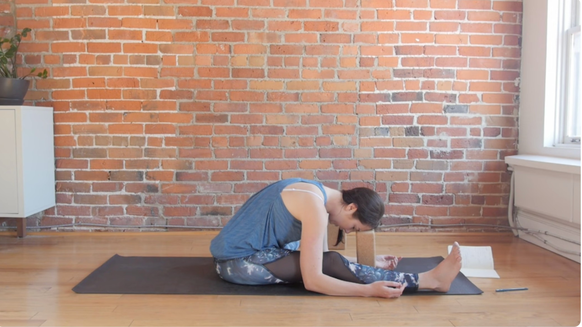 Woman sitting on a mat in Half Butterfly Pose and leaning forward with her forehead or Third Eye chakra or Ajna on a yoga block