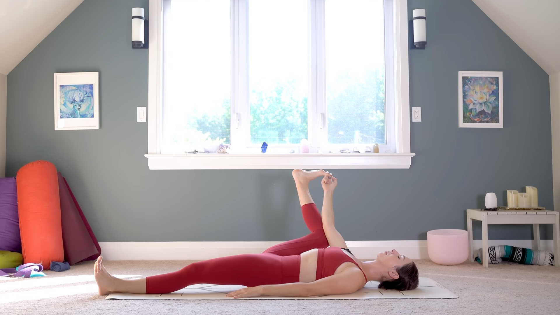 Yoga With Kassandra lying on her back with her hand reaching for her right foot in Ardha Ananda Balasana