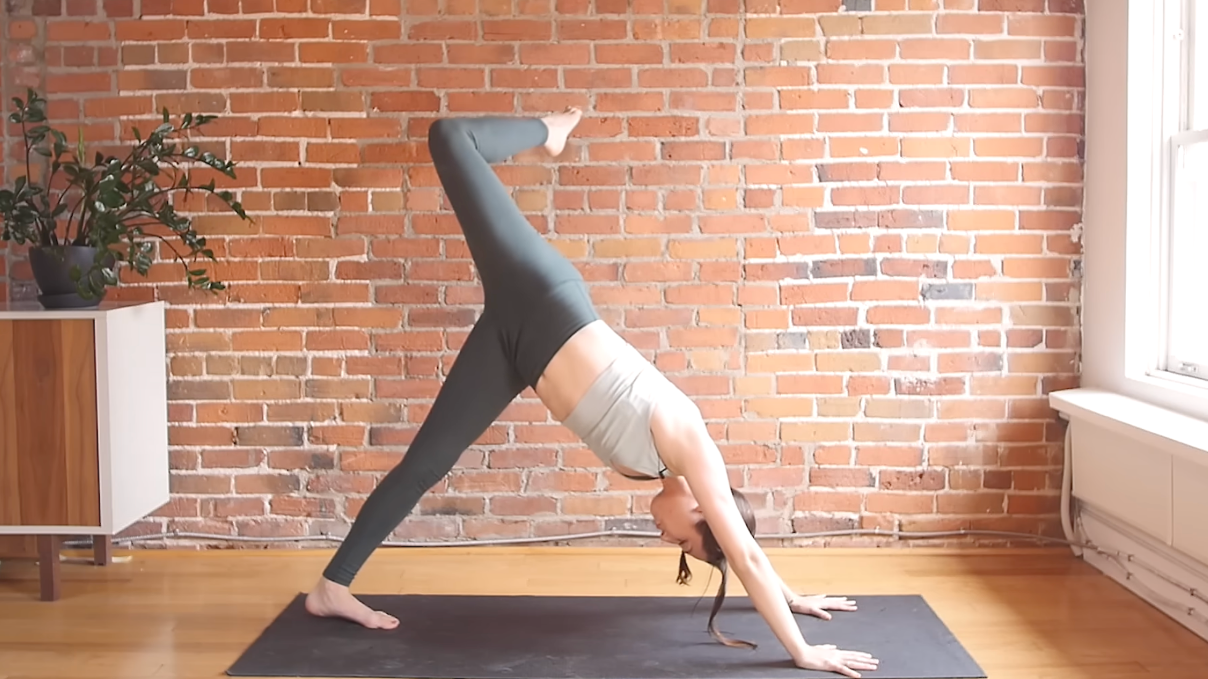 woman in yoga clothes in Three-Legged Dog pose with both hands and left foot on ground and hips raised up, with right leg lifted above hips and knee at 90-degree angle on gray mat in front of brick wall background