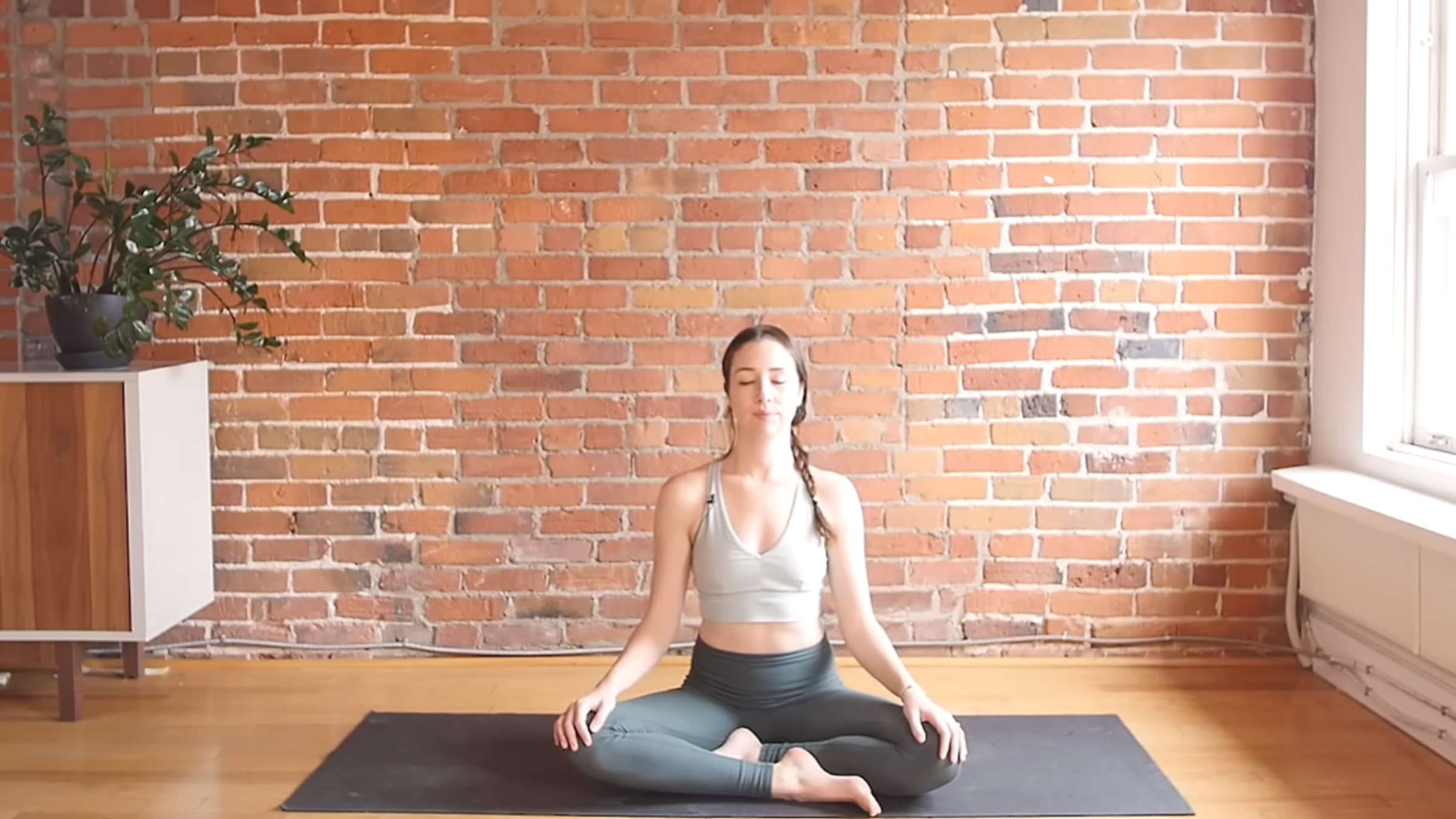 woman in yoga clothes seated on yoga mat with knees bent and hands on knees in front of brick wall background