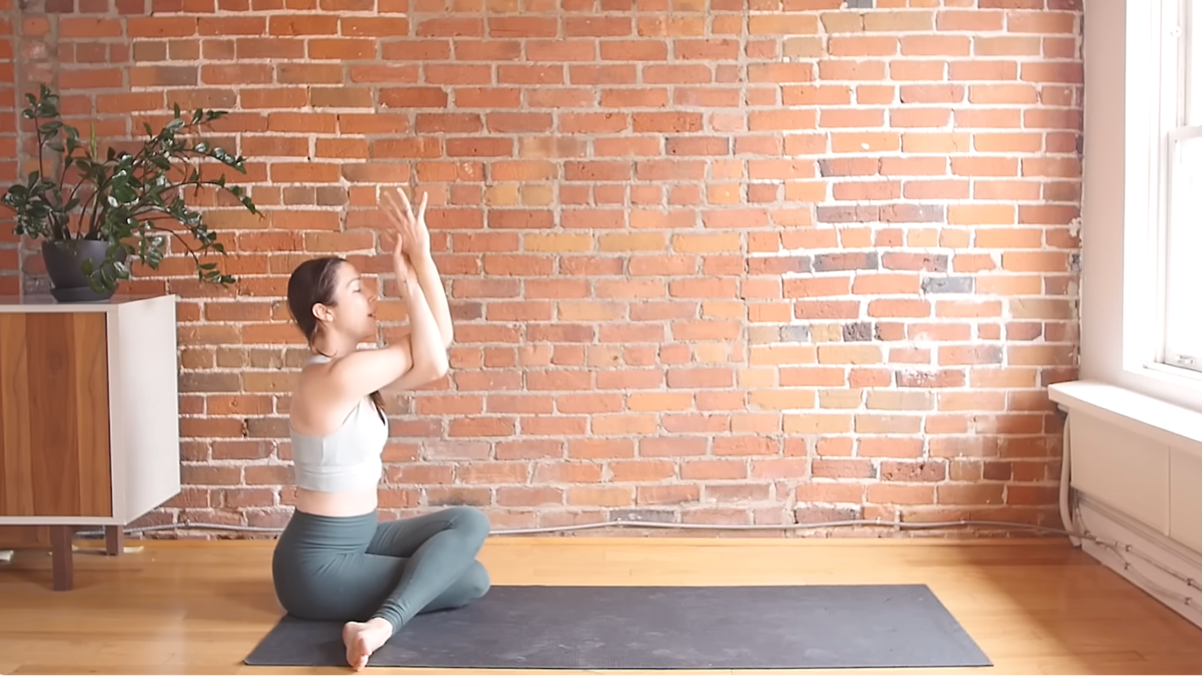 woman with legs in Cow Face Pose with left leg bent and crossed over right leg with both feet toward back corners of mat and arms twisted together in Eagle Arms pose with brick wall background