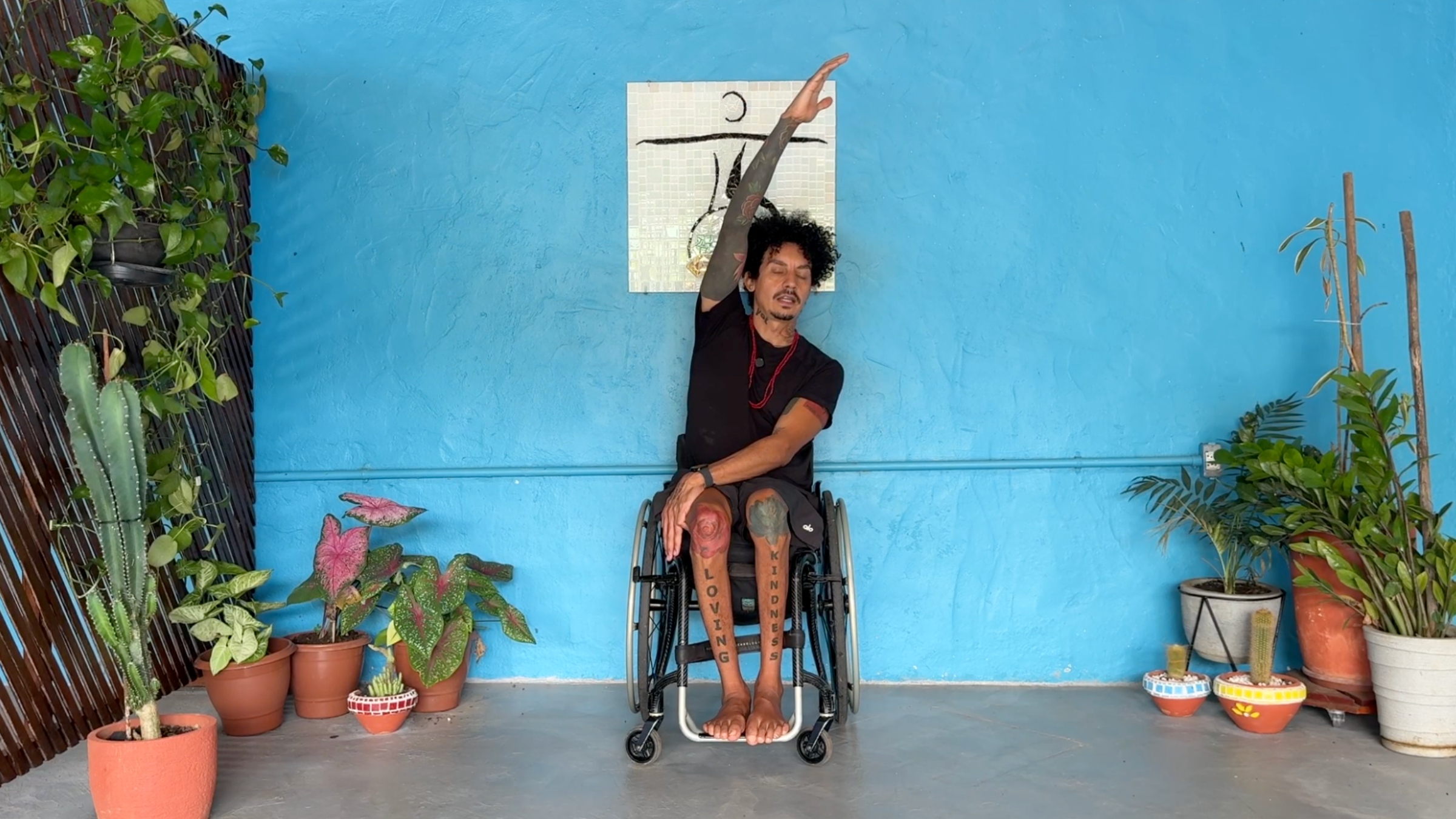 man seated in wheelchair facing the camera in Seated Side Bend with left arm crossed to outside of right knee and right arm extended overhead and toward the left side