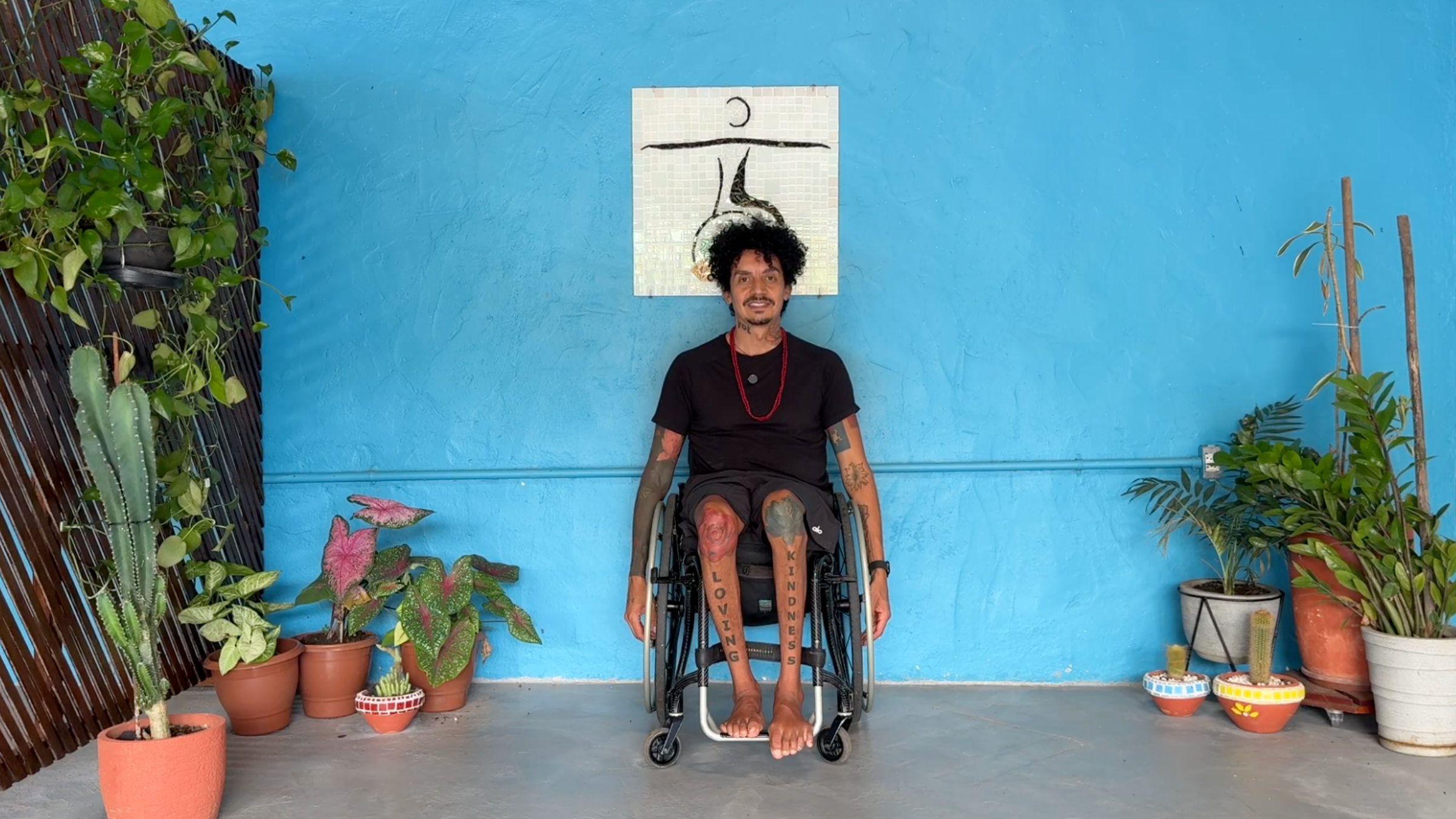 man in wheelchair in seated Mountain Pose sitting up tall with arms extended down toward the floor in front of blue wall with plants around the room