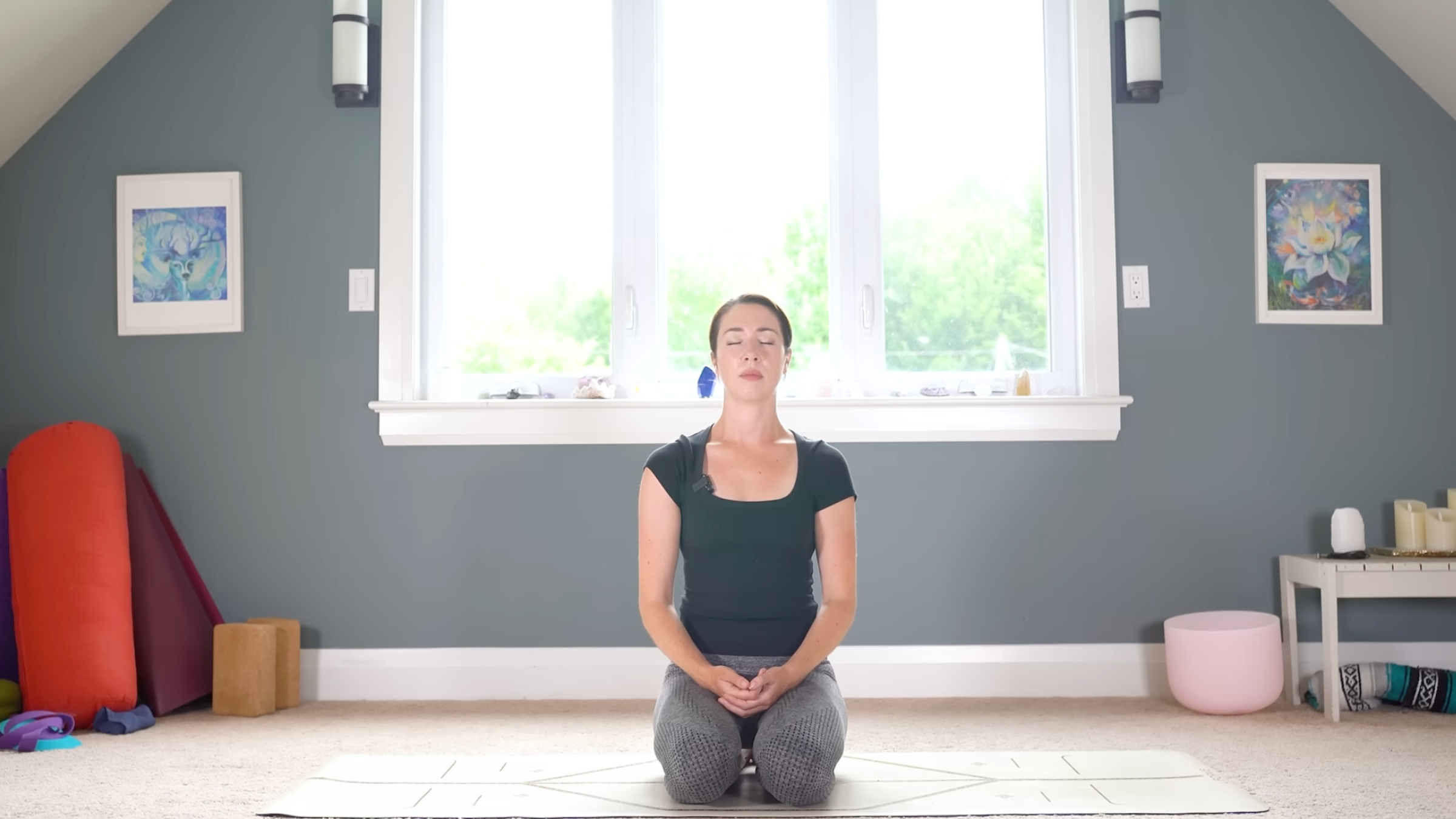 woman in yoga clothes kneeling on yoga mat on carpeted floor in home in resting position with fingers interlaced and resting in lap and eyes closed