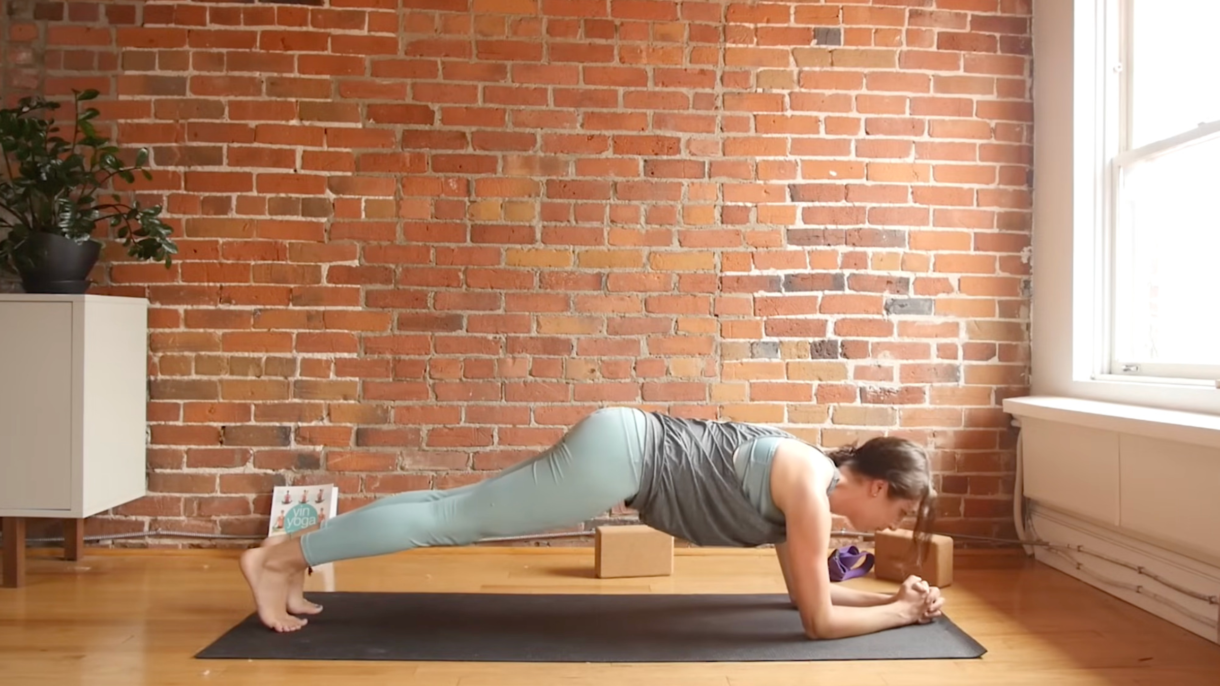 woman in yoga clothes on yoga mat in Forearm Plank pose with forearms on mat, fingers clasped, and body in straight line