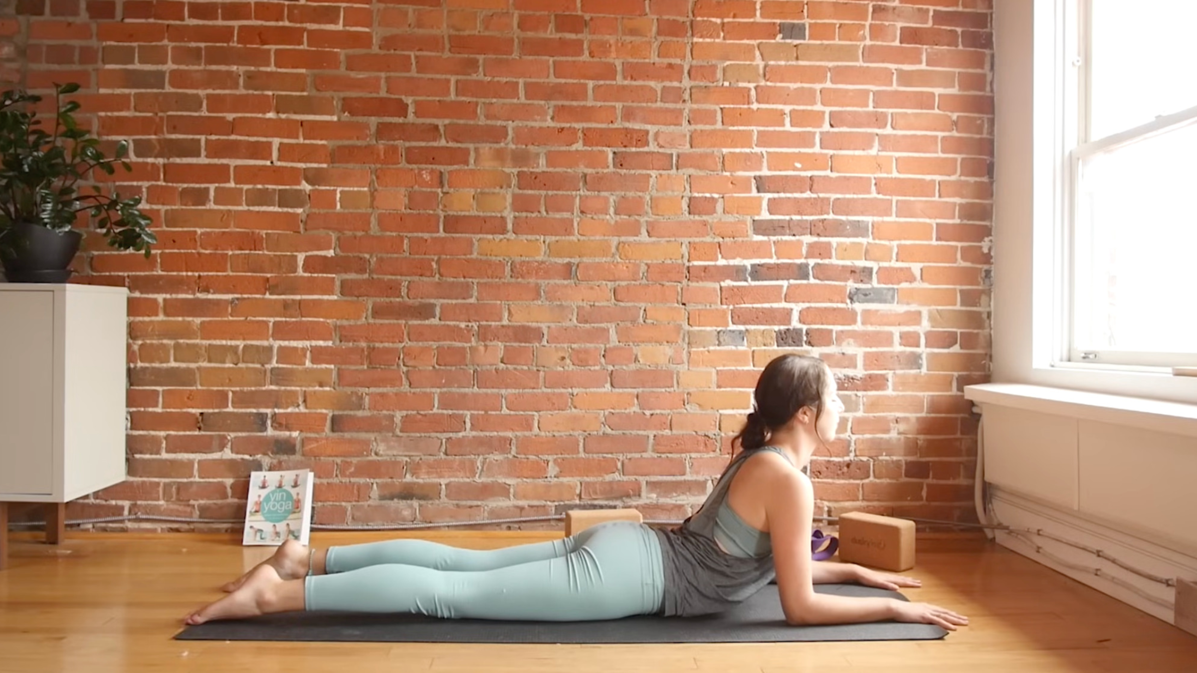 woman in yoga clothes lying face forward on yoga mat in Sphinx Pose, with shoulders stacked over elbows, forearms and hands on the floor, legs on the ground, and chest and head lifted, head facing forward