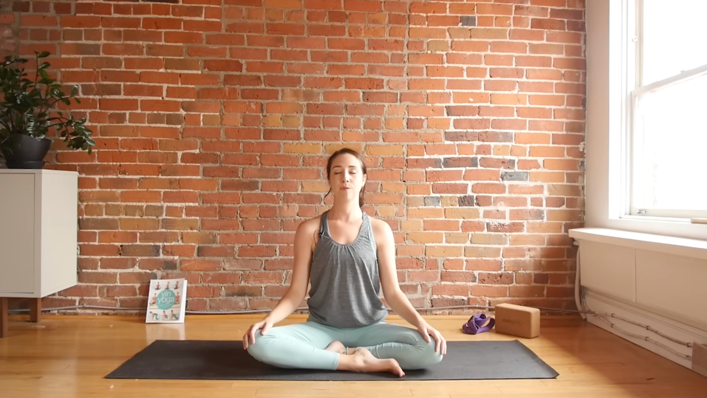 woman in yoga clothes sitting on yoga mat in Closing Seat, with legs crossed, back straight, eyes closed, and hands resting on knees