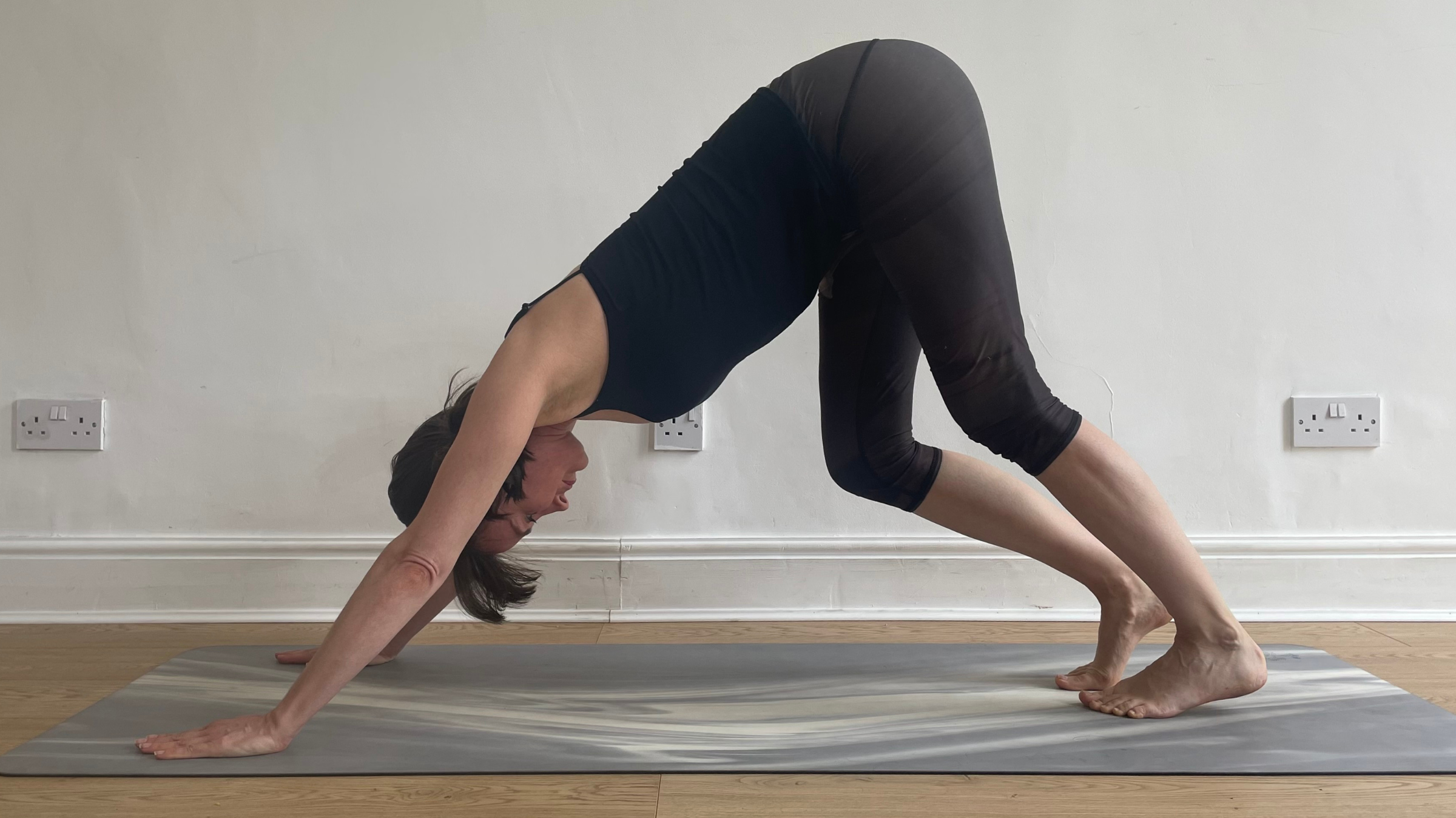 Amber Burke in Downward-Facing Dog pose, demonstrating a calf stretch by keeping her hips high while bending one knee deeply and pressing the opposite straight leg's heel toward the mat.