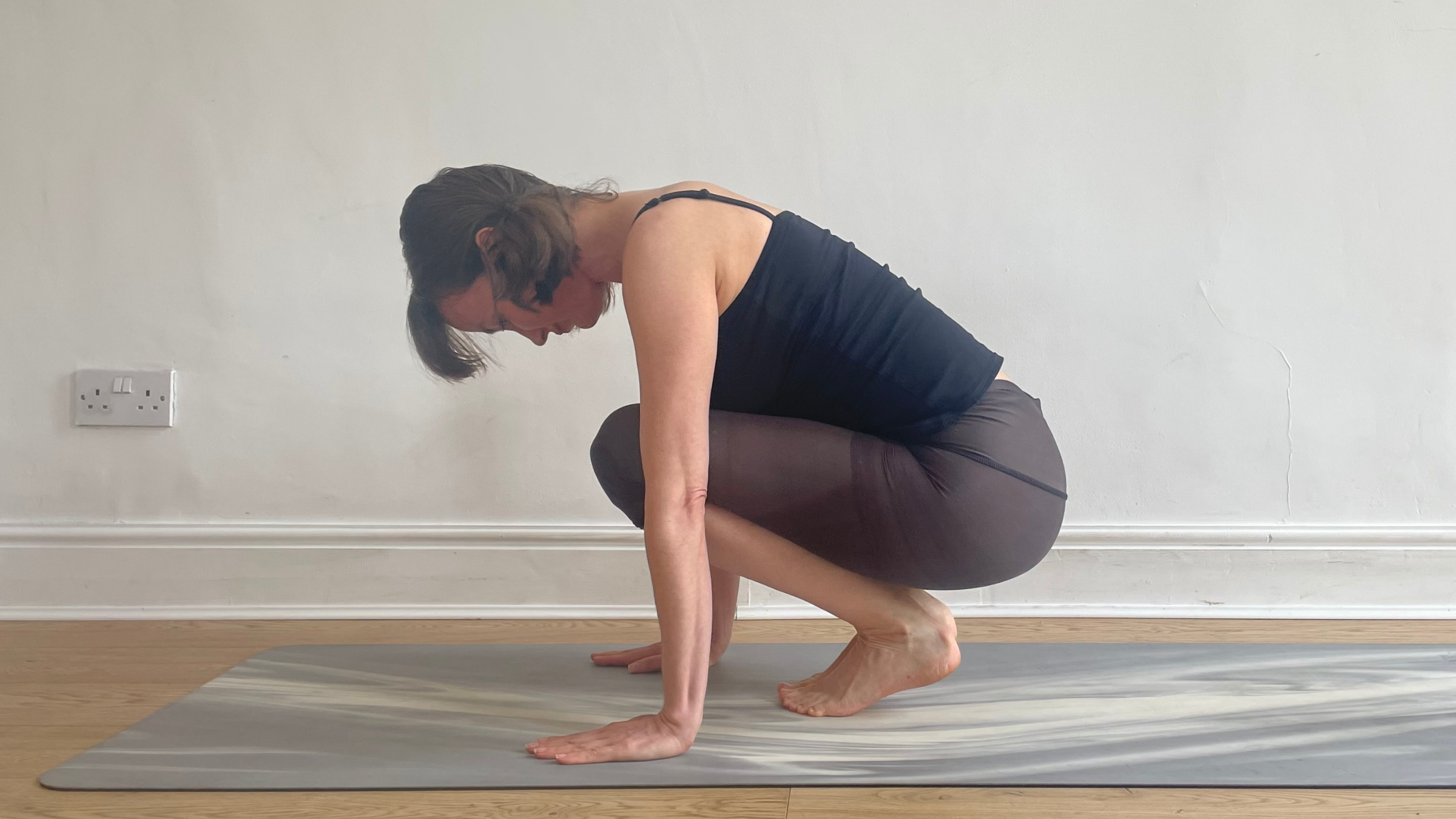 Amber Burke crouching on a yoga mat in a tight, rounded ball shape, balancing on the balls of her feet with her heels hovering just off the ground and her chest resting against her thighs.