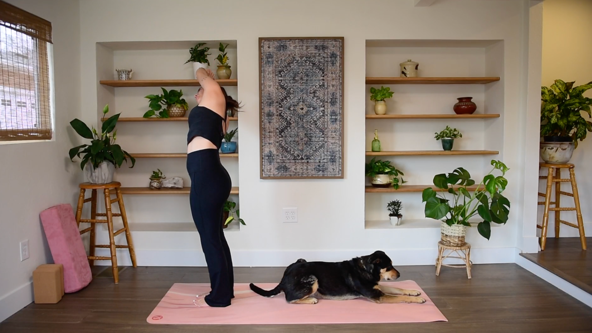 Caitlin K'eli standing on a yoga mat with arms clasped overhead, gently leaning her torso to the side in a Standing Side Bend.