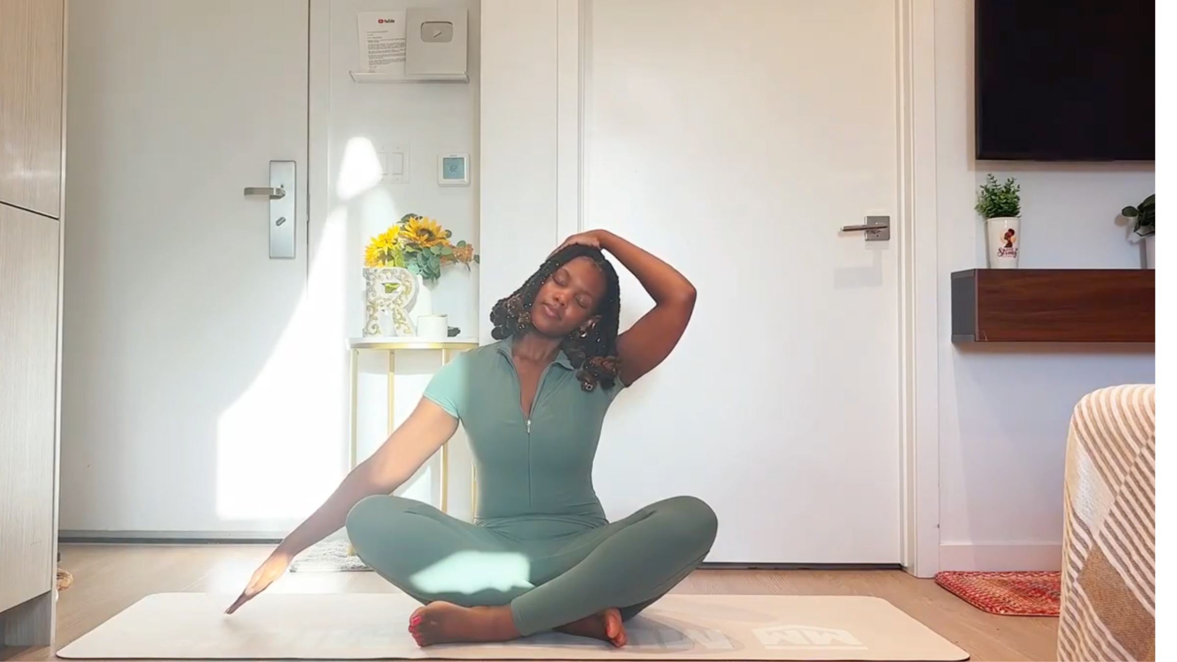 Ramoni Overton sitting cross-legged on a yoga mat, gently tilting head toward shoulder with one hand resting on head to stretch the neck.