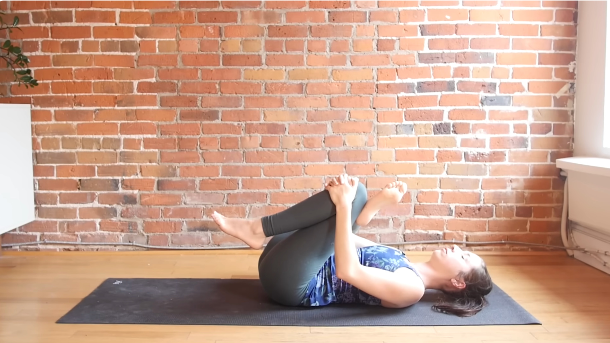 woman in Reclined Pigeon pose lying on back on mat with right ankle crossed over left knee, both knees bent, hands clasped around left knee and pulling it toward chest