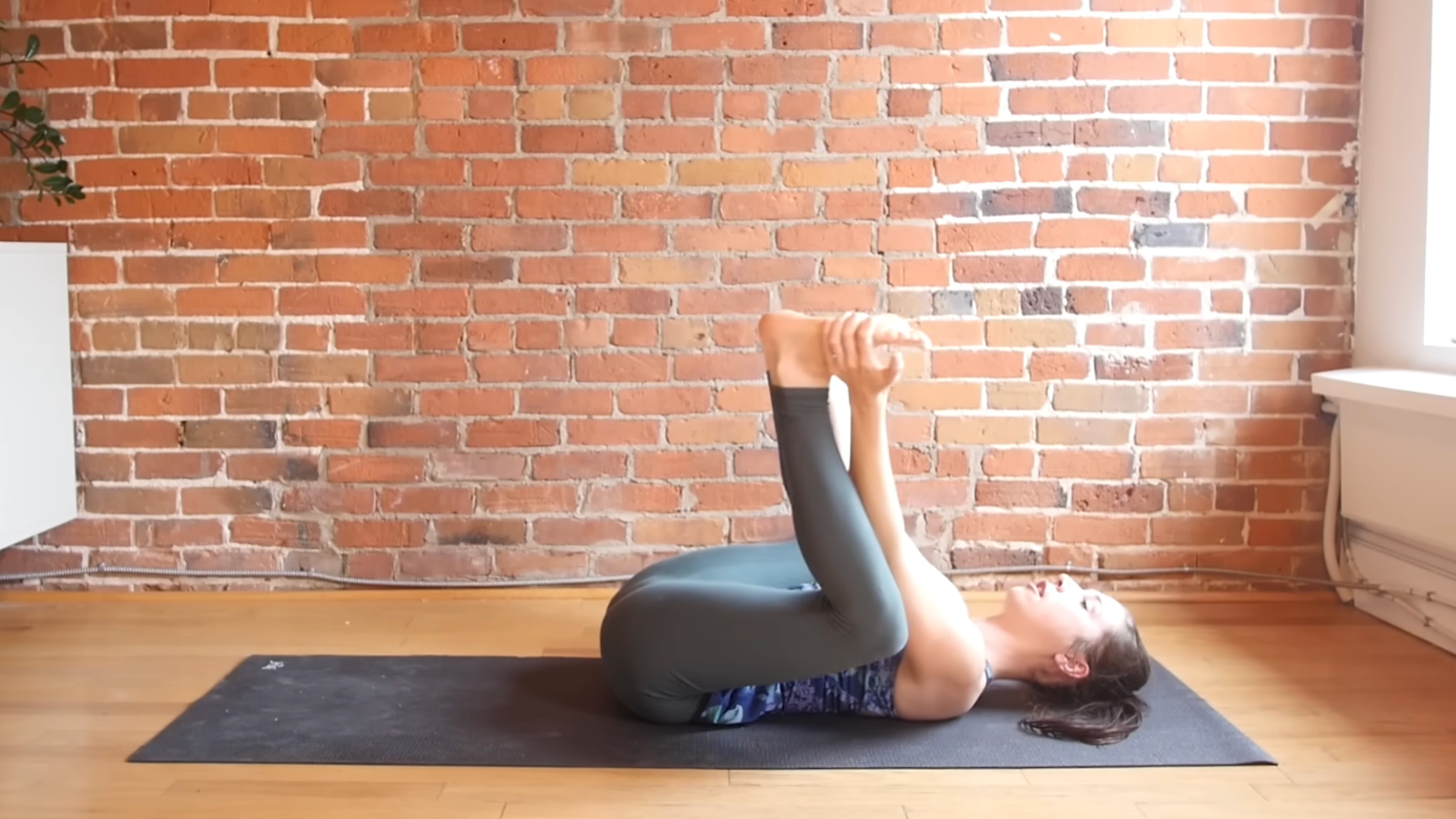 woman in yoga clothes on yoga mat in Happy Baby pose with knees bent, feet flat toward the ceiling, and hands grabbing outside of feet