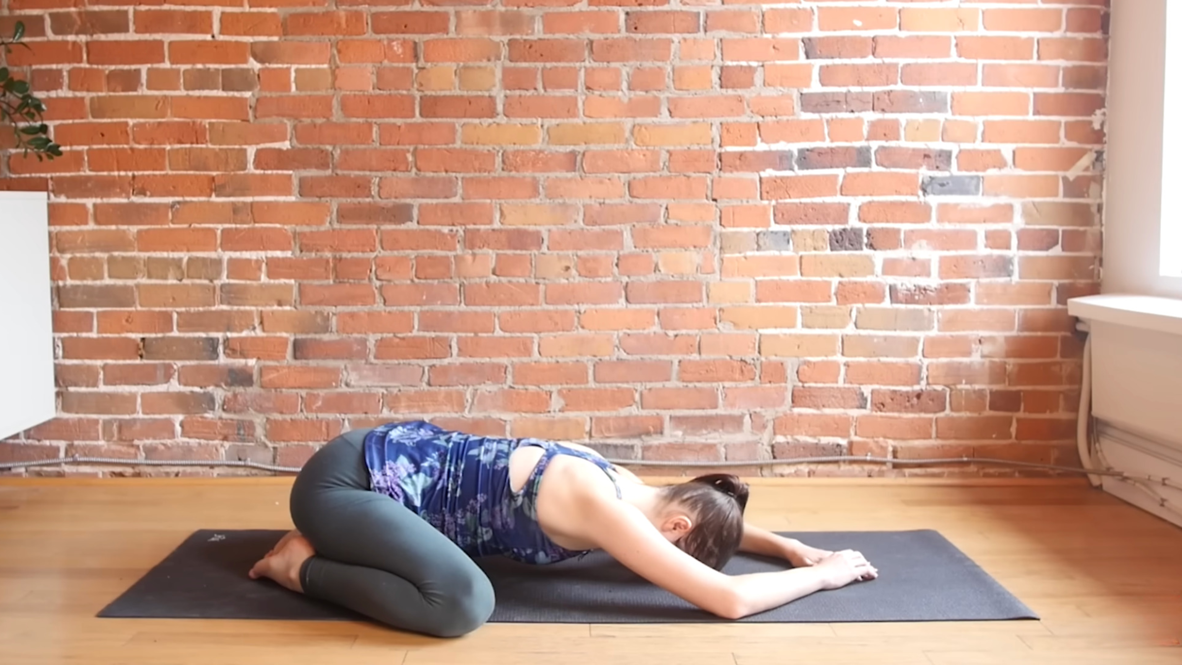woman in yoga clothes in Child's Pose with big toes touching, knees out past edges of mat, sitting back on heels and forehead and arm resting on the mat